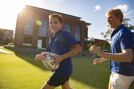 A boy in a blue polo shirt and navy shorts running with a rugby ball being chased by another boy in a blue shirt on a green field in front of a large brick building.